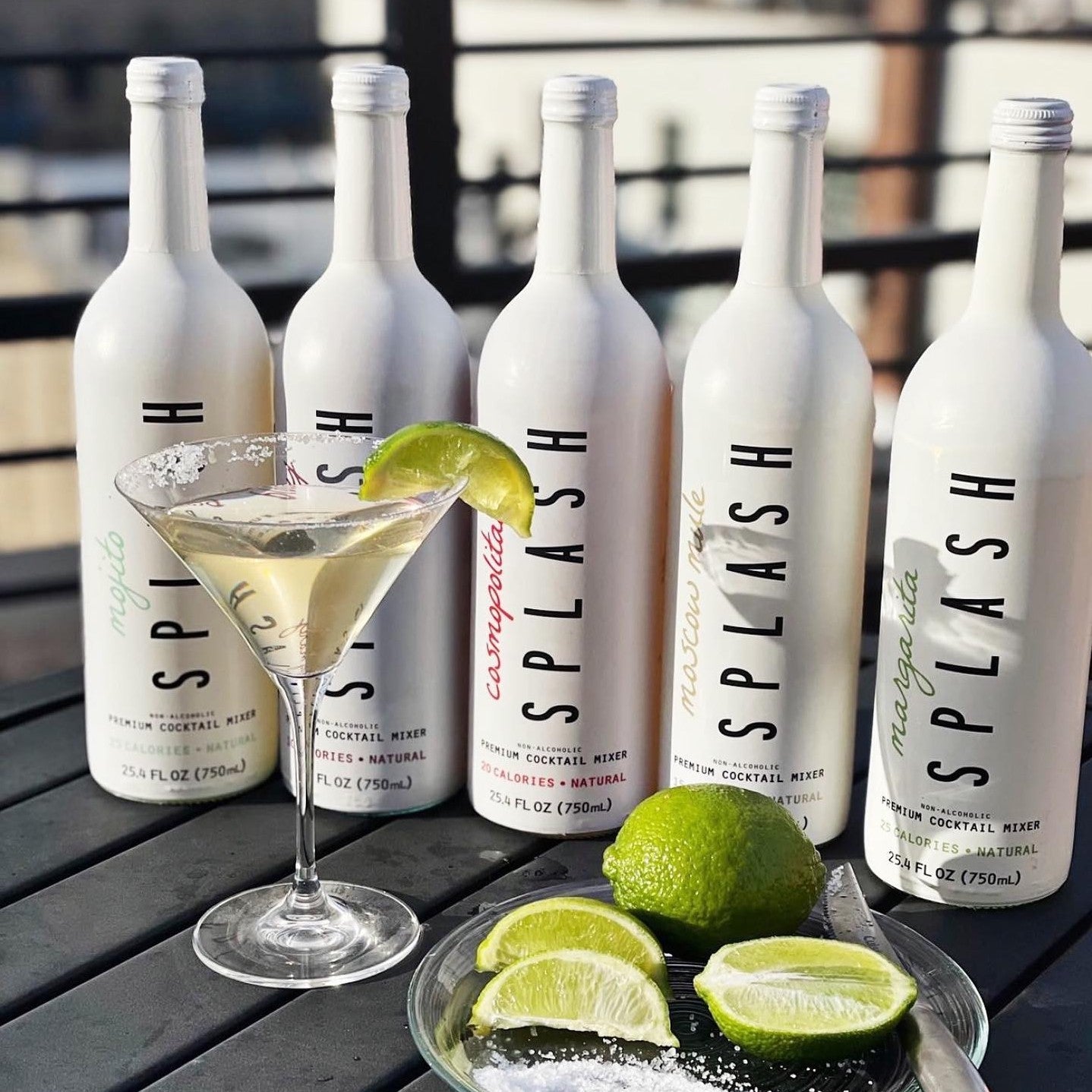 All five flavors of Splash Mixers on a table outside, next to a cocktail in a martini glass, garnished with lime.